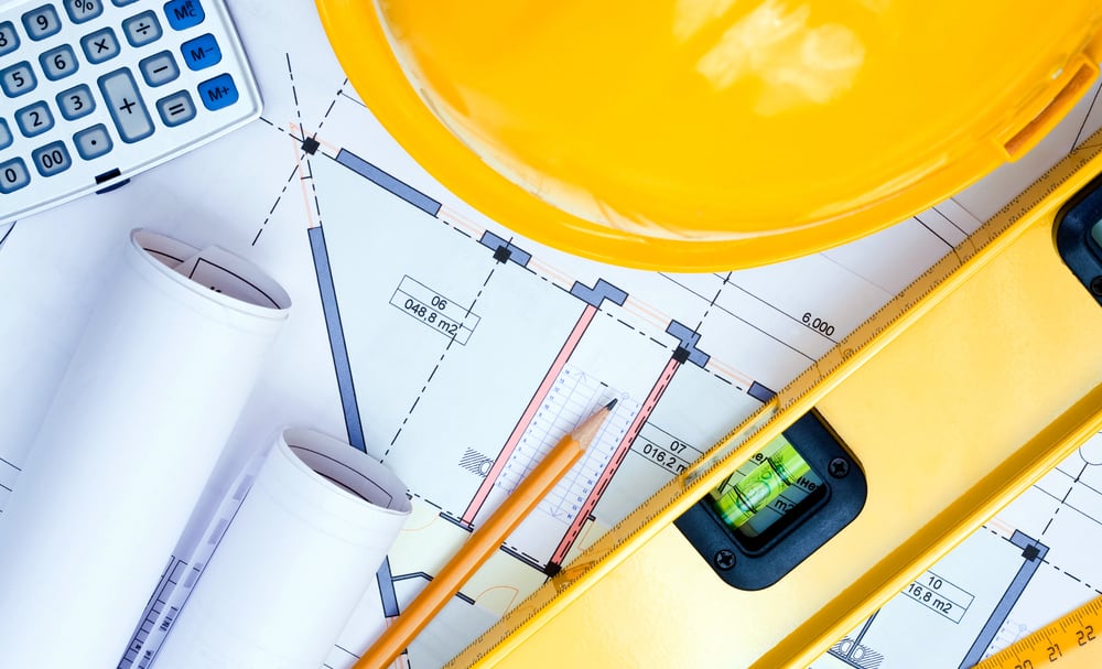  A table with a job plan underneath a construction helmet, level, pencil, and calculator to represent the best customer financing for contractors